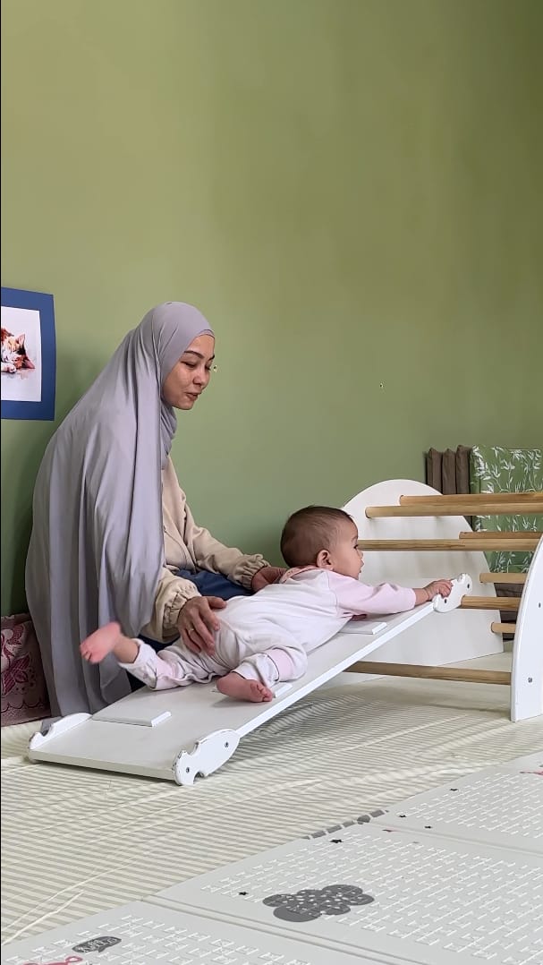 Pikler qiyaligida harakatlanayotgan chaqaloqni qo'llab turgan ota-ona